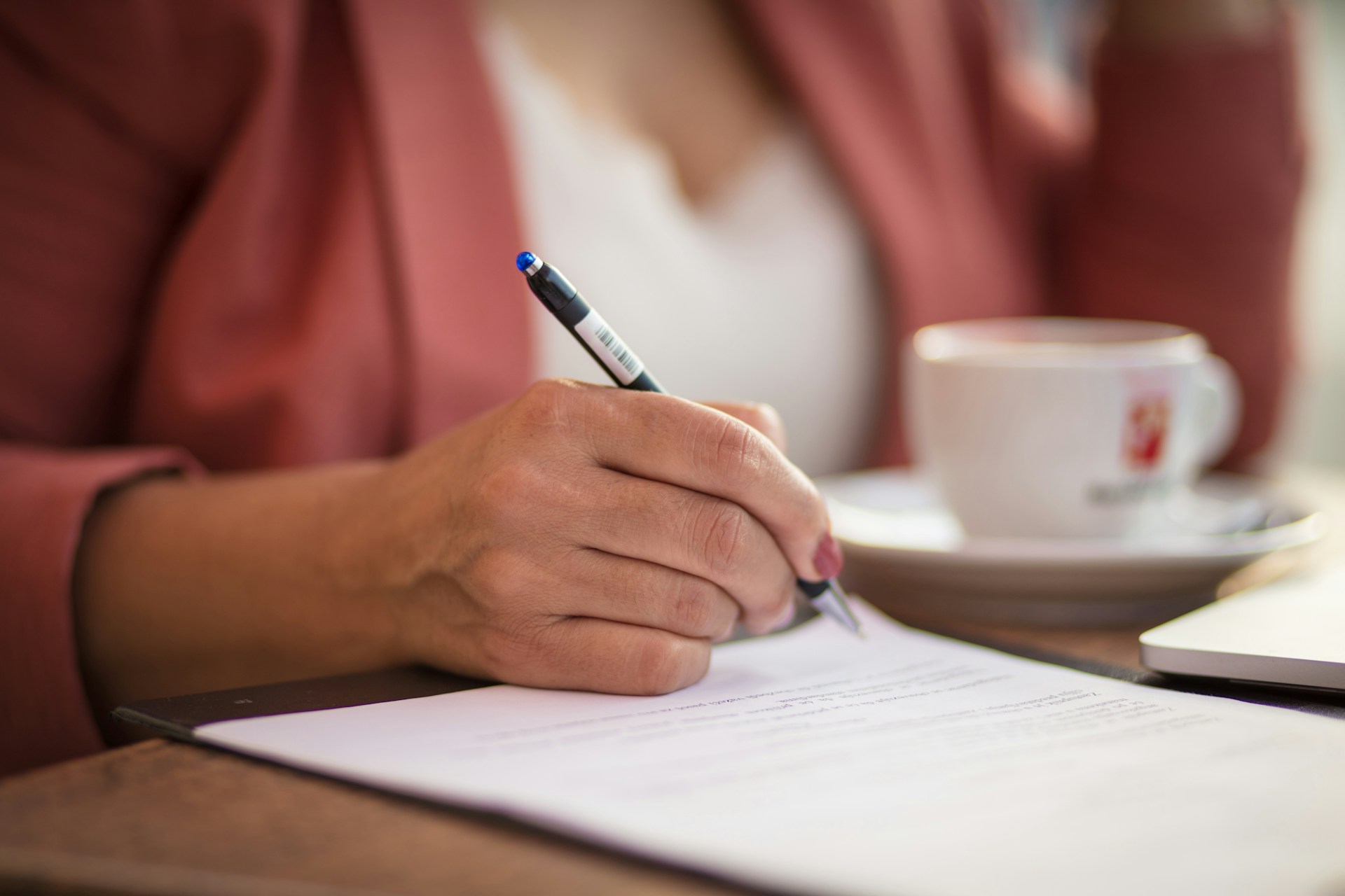 A woman, shown from the shoulders down, writes out her will with a pen on paper next to a cup and saucer set on the table in front of her, representing the validity of draft wills in BC.