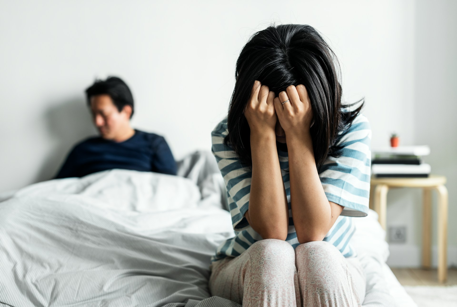 A woman sits on the end of the bed with her face buried in her hands, and her blurred husband is sitting up at the top of the bed under the covers, representing intimate partner violence in BC family law.